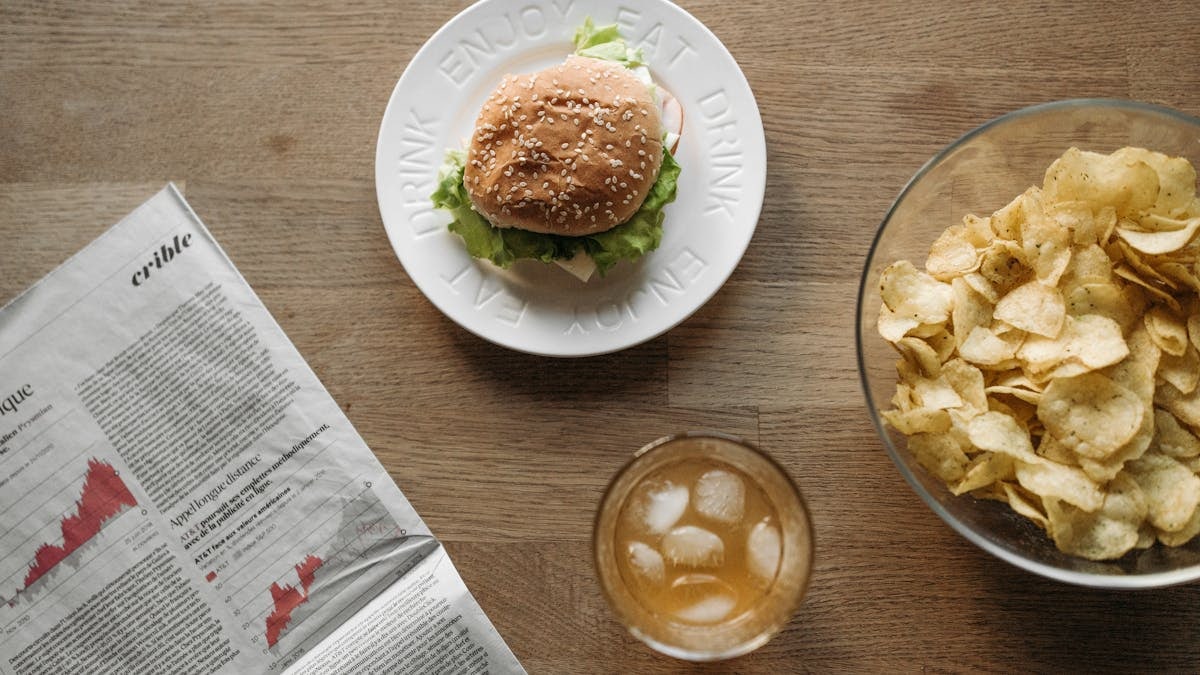 Overhead shot of a burger, chips, and drink on a wooden table with a newspaper.