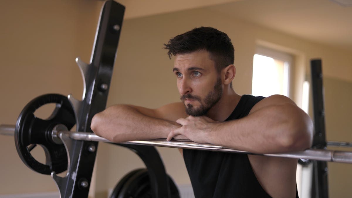Concentrated muscular bearded sportsman in black activewear leaning on metal barbell and looking away while resting during training in modern gym